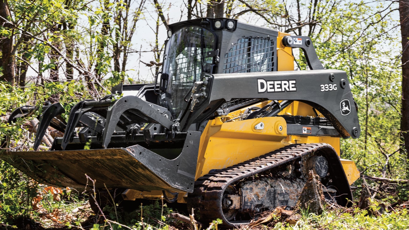 A 333G Compact Track Loader with grapple attachment in front of trees. 